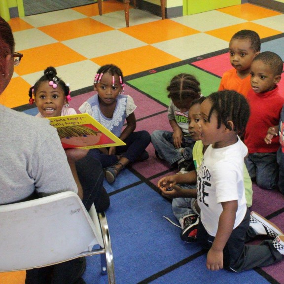 children sitting on a floor listening to a teacher read to them.