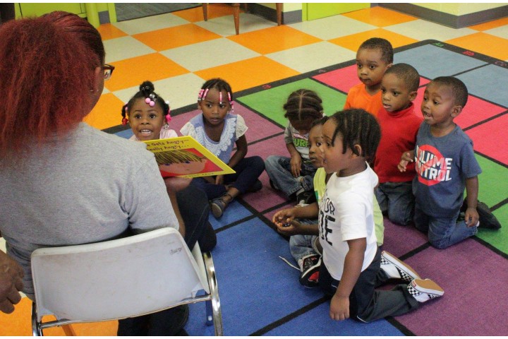 children sitting on a floor listening to a teacher read to them.